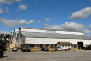 Large industrial warehouse with a metal exterior, several parked vehicles in front, and a small white shed beside the building under a partly cloudy sky.