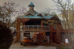 A brick building under construction with a green roof, a gazebo-like upper level, and an excavator and worker in front. Some trees and traffic cones are visible nearby.