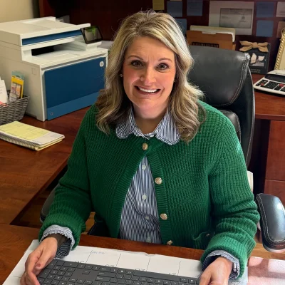 Woman with blonde hair wearing a green cardigan sits at a desk with a keyboard, office supplies, and printer visible in the background.