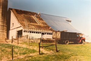 Workers install a new metal roof on a large barn, with a red truck parked beside it and a silo visible on the left.