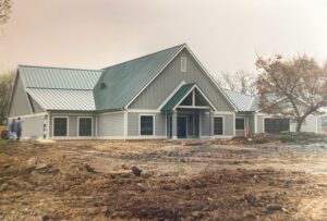 A newly constructed building with gray siding and a green metal roof, surrounded by dirt and some construction materials; two people stand near the left side.