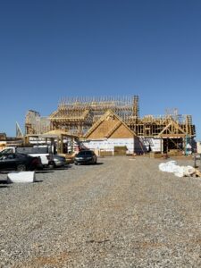A wooden-framed building under construction, with construction materials and parked cars on a gravel lot under a clear blue sky.
