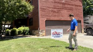 A man in a blue shirt stands near a yard sign for Henley Roofing Company in front of a brown house with a closed garage door.