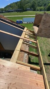 View from a rooftop showing construction work with exposed wooden beams and planks; tools and equipment are visible below on the grass.