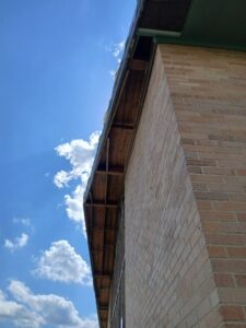 View looking up at the edge of a brick building with exposed wooden eaves against a blue sky with scattered clouds.