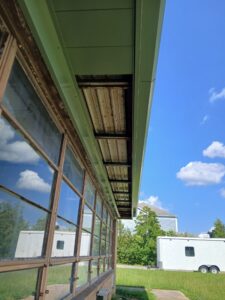 A close-up view of a building with upper soffit panels partially missing, exposing the roof structure; clear sky and trailers are visible in the background.