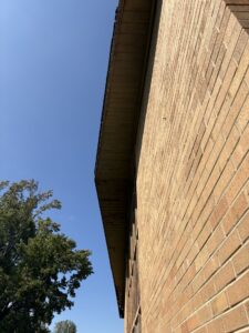 A brick building is shown from a low-angle perspective against a clear blue sky, with part of a leafy tree visible on the left side.