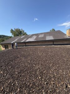 Two people stand near a large sloped roof with visible repairs or discoloration; the foreground shows a flat, gravel-covered surface under a clear sky.