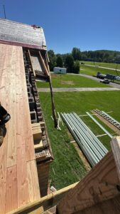 View from a roof under construction, showing exposed wooden beams, stacked metal roofing sheets, ladders, and grassy area with vehicles in the background.
