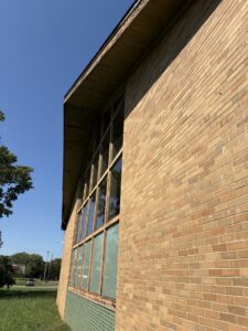 Close-up of a brick building with large vertical windows under a slanted roof, taken on a sunny day with a clear blue sky.